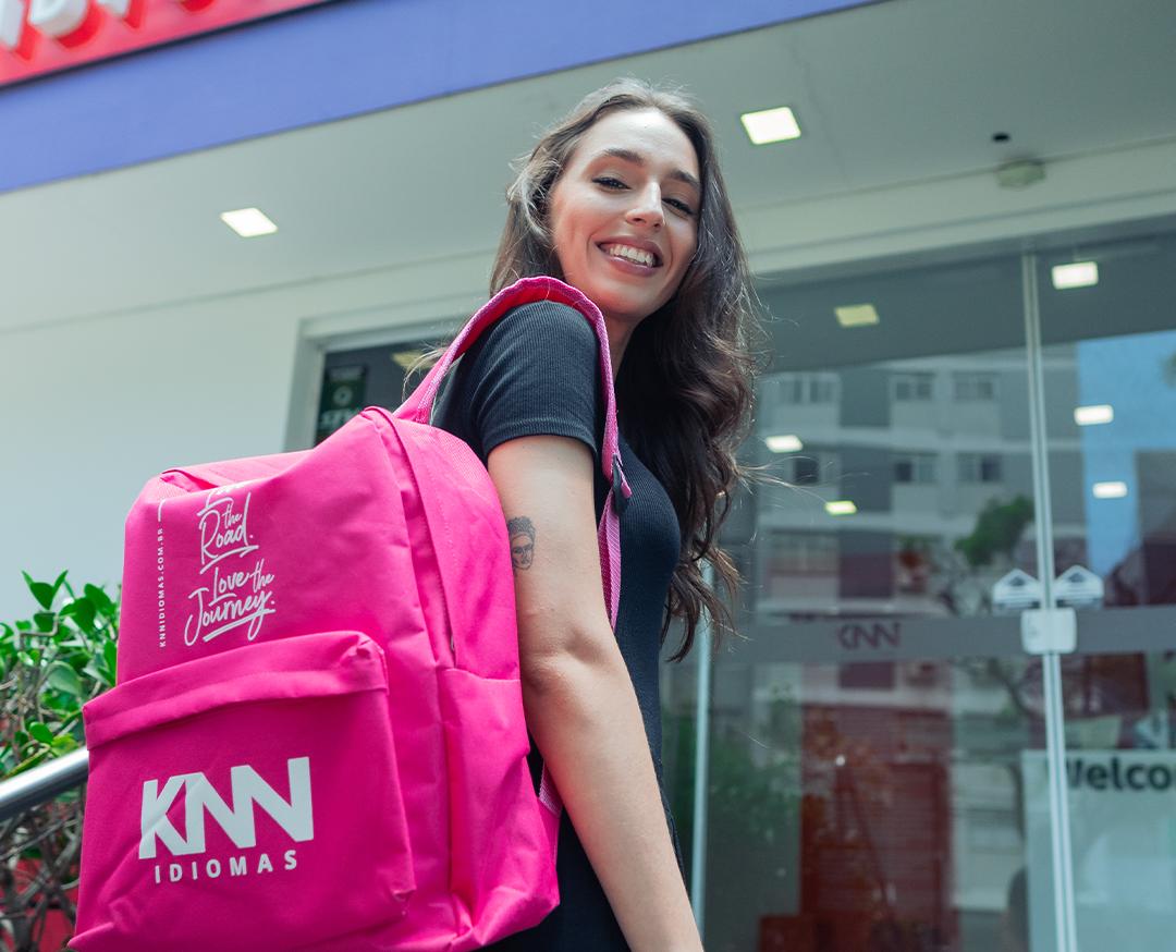 Menina na entrada de uma unidade da KNN com um mochila rosa nas costas.